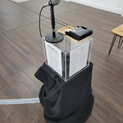 a microphone and a tape recorder atop a clear acrylic plinth with artwork info on the sides. A black cloth is draped underneath covering the rest of the plinth