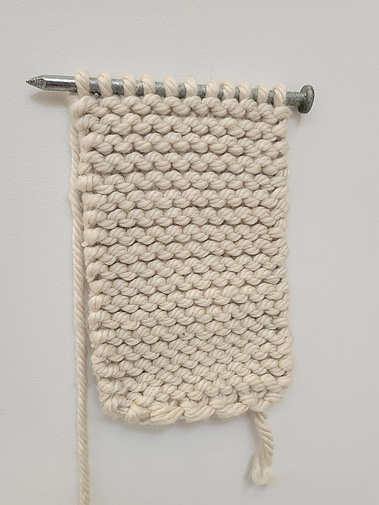 detail photo of incomplete knit scarf. Hanging off an oversized steel nail (knitting needle).