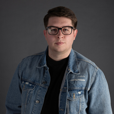 A man, wearing large black glasses, a black t-shirt and a jean jacket stands in front of a gray background.