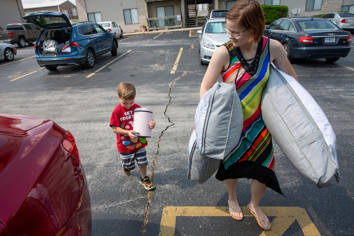A woman is carrying a pair of pillows. Behind her, a young child carries a small trash can.
