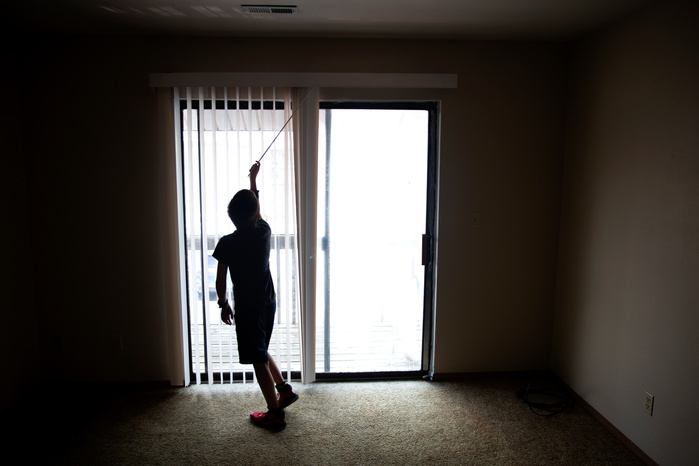 A boy stands in silhouette and opens the blinds covering a glass door.