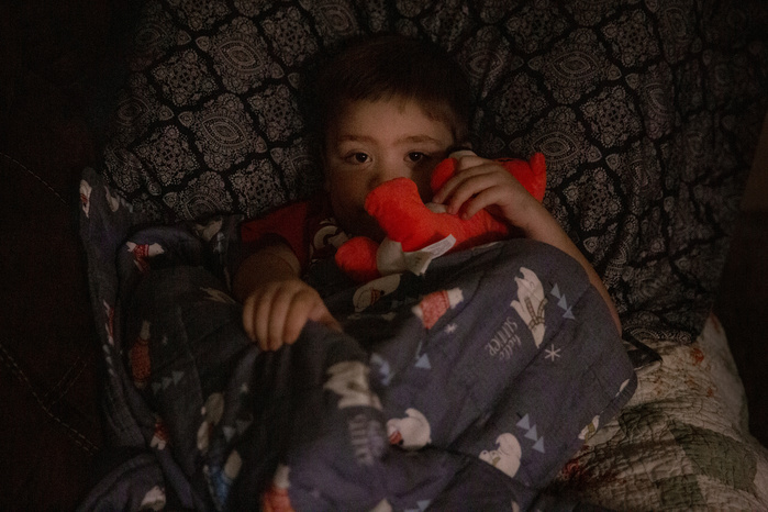 A young boy looks into the camera. His face is partially obscured by a red teddy bear he is holding. His other hand is grasping a blue blanket decorated with polar bears.