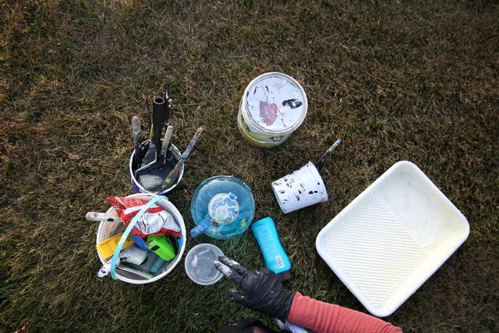 A hand reaches for a pile of paint and brushes resting on the grass.