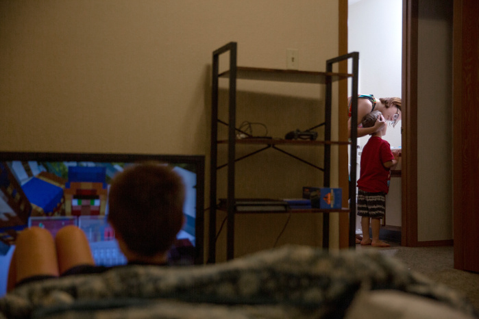 In the foreground, a young boy plays a video game on a television screen. In the background, a woman helps another young boy brush his teeth.