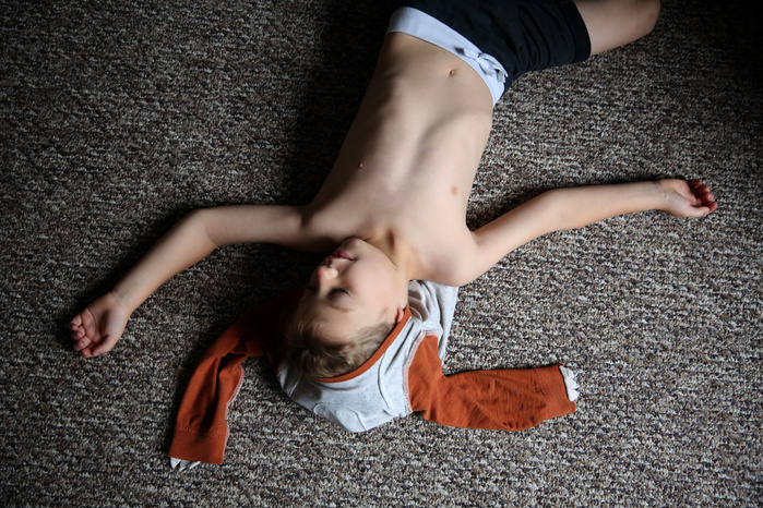 A young boy lays out on the ground, upside down. His arms are spread out like a cross. His shirt is up and around his head, the sleeves also spread out like a cross.