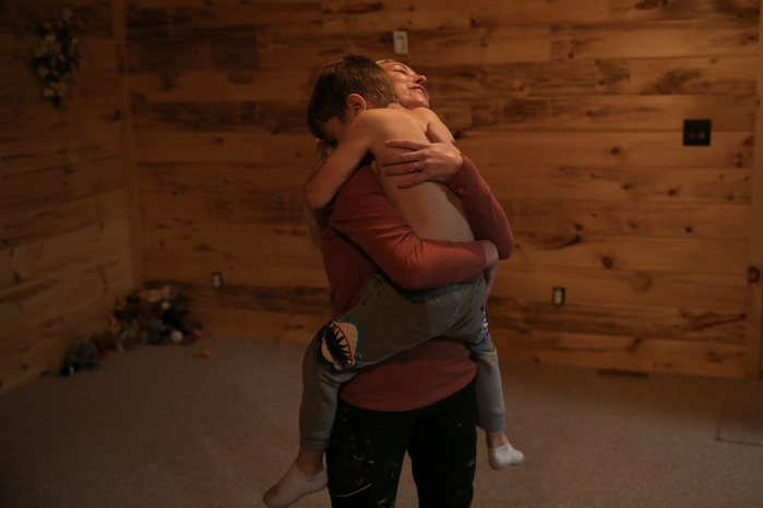A woman lifts a child off the ground in a hug. Behind them, the walls are wood and a pile of toys sits in a corner.