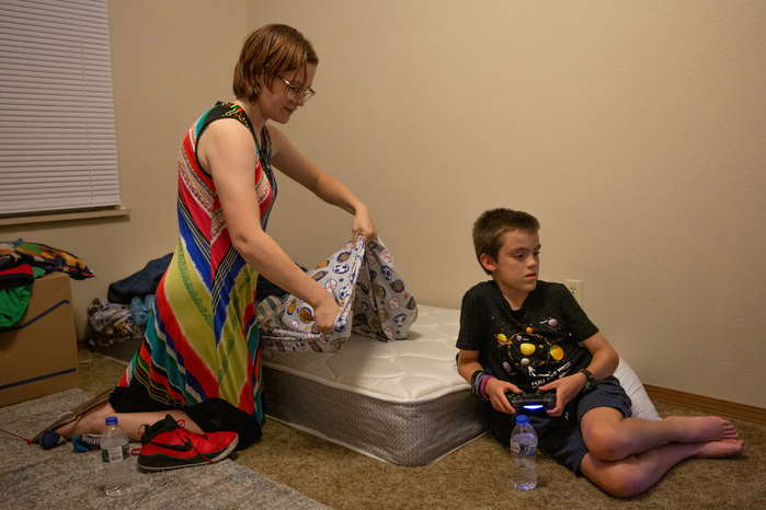 A woman lays a sheet onto a mattress. The mattress is resting on the floor. Sitting near the mattress is a young boy holding a video game controller.