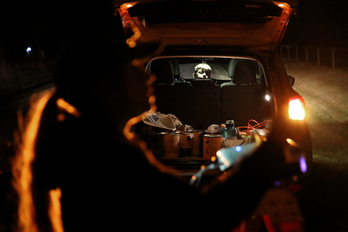 In the foreground, a silhouetted woman holds a projector. In the background is a car. Inside the car, a young boy looks at the woman, lit by the interior car light.