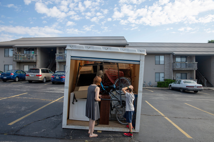 A woman and boy stand in a parking lot, looking into a storage pod filled with boxes and furniture.