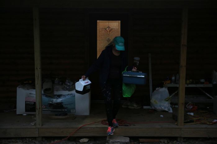 A woman holding a lunchbox and a toolbox steps off a short porch. Her face is obscured by a teal hat.