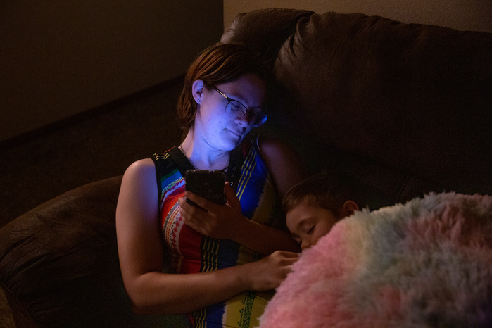 A woman holds a small boy in her lap, looking at him while he sleeps. Her face is lit blue by a cell phone.