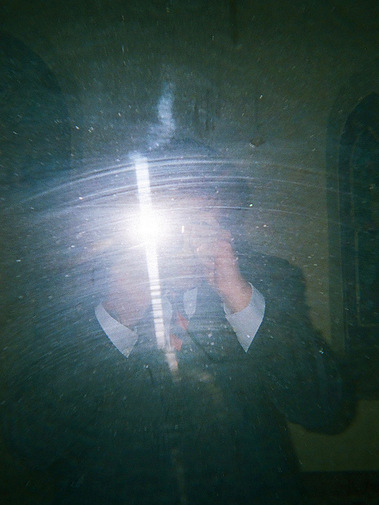 A white man wearing a dark three piece suit and red and navy tie takes a picture in a dirty mirror. The flash of the camera is obstructing his face. 