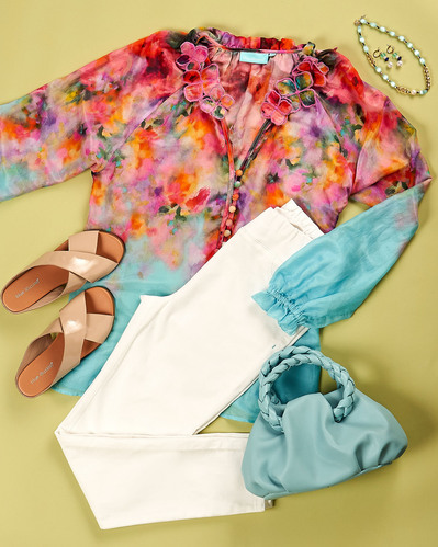 a flatlay of floral blouse, white pants and accessories on a green background