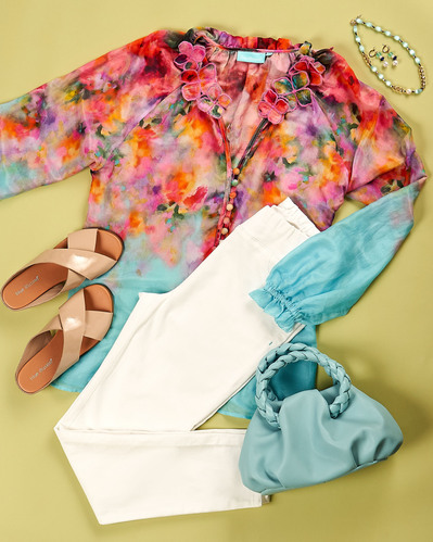 a flatlay of floral blouse, white pants and accessories on a green background