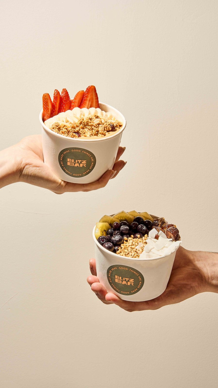 Two acai bowls with fresh toppings held against a neutral wall, featuring strawberries and blueberries