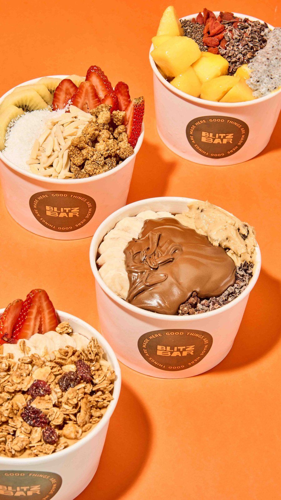 Trio of acai bowls topped with granola, strawberries, mango, and Nutella against vibrant orange background