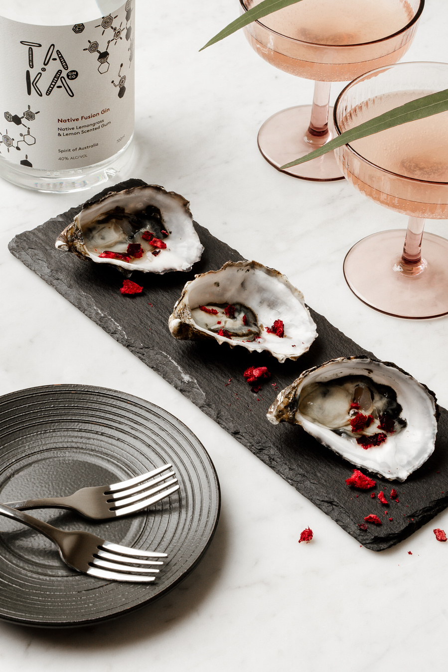 oysters on a dark plate with two pink cocktails next to it and a bottle of alcohol
