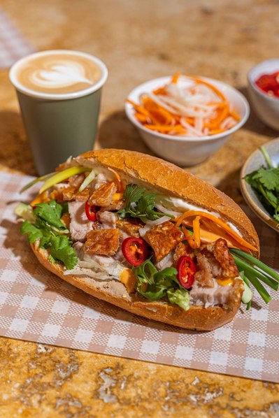 a big banh mi sandwich lying on the table with a cup of coffee and sides next to it