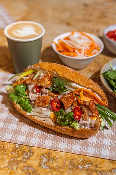 a big banh mi sandwich lying on the table with a cup of coffee and sides next to it