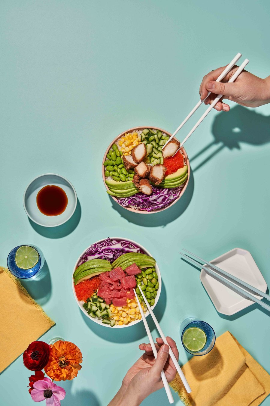 Hands using chopsticks to enjoy vibrant poke bowls on a pastel teal table setting with fresh flowers and drinks