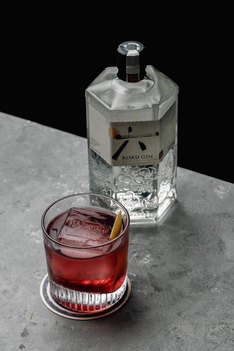 Negroni cocktail with a lemon twist next to a stylish gin bottle on a gray stone surface