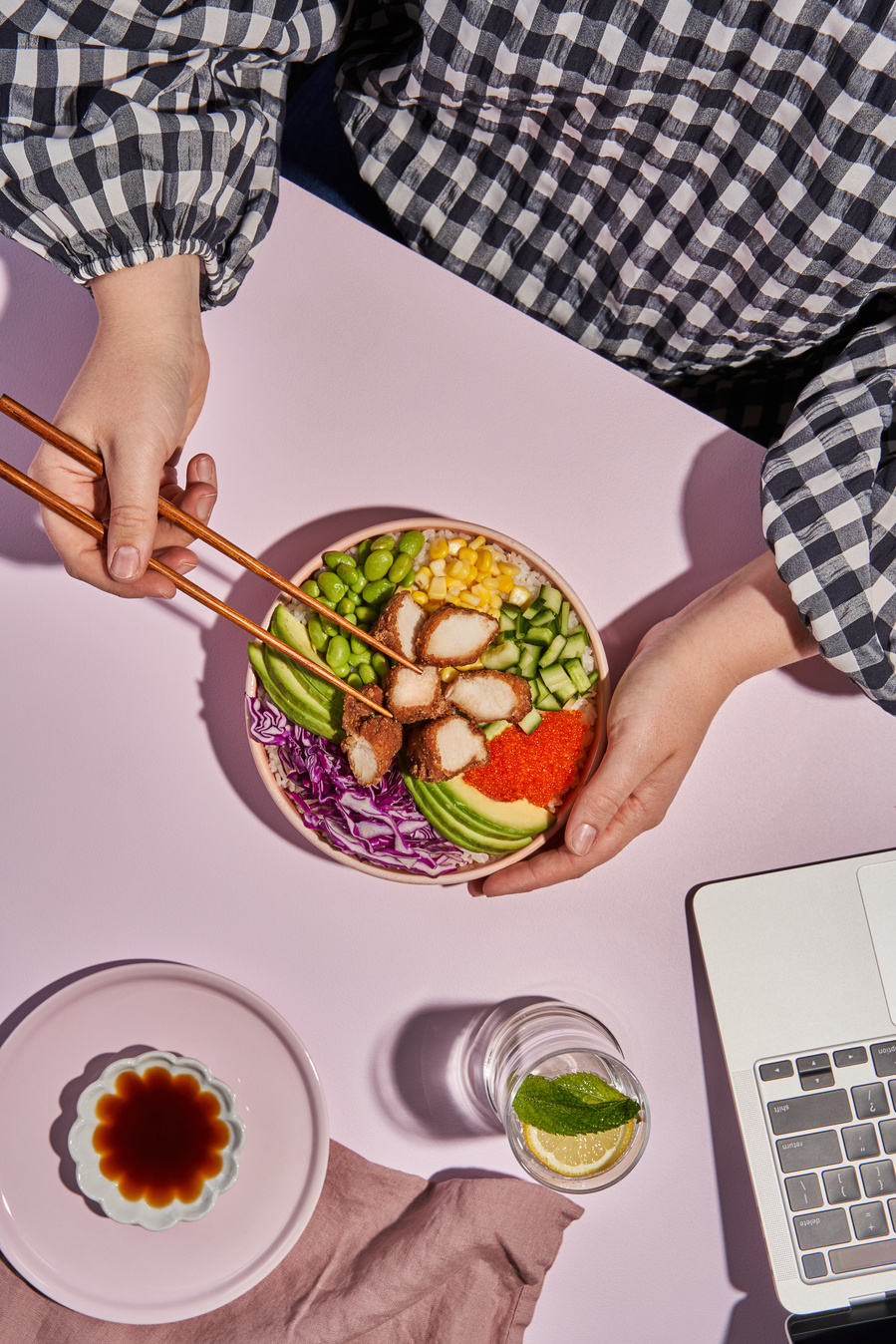 hands holding a poke bowl on a pink table with laptop, a drink and soy sauce in the frame