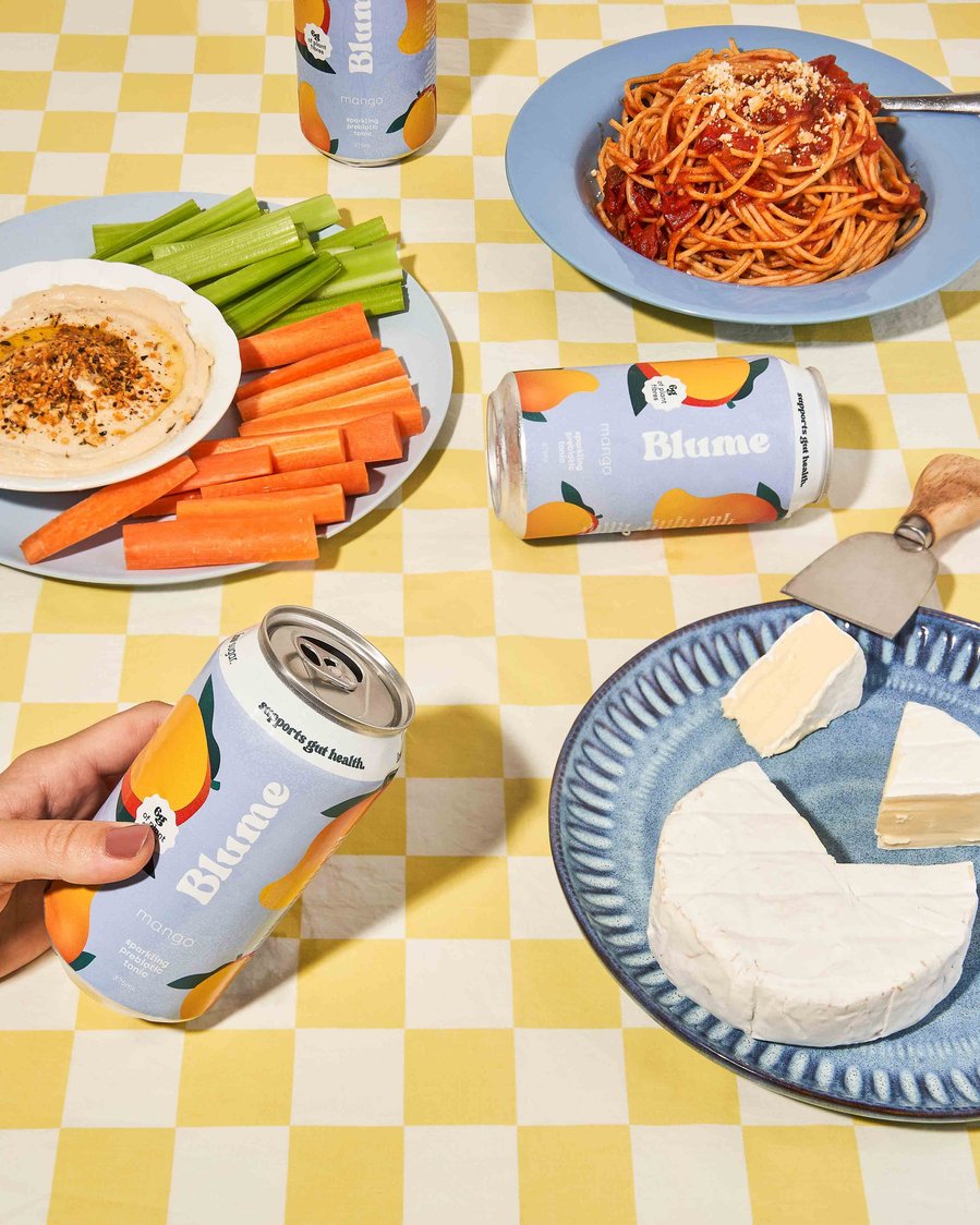 a colourful food spread with brie, pasta, fresh veggies and a hand holding a kombucha 