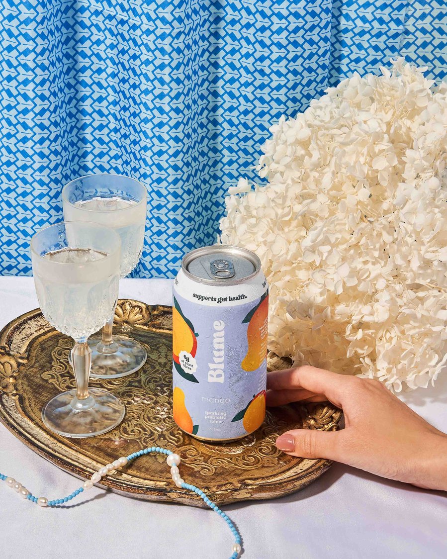 a hand holding a blue kombucha with glasses and flowers in the background