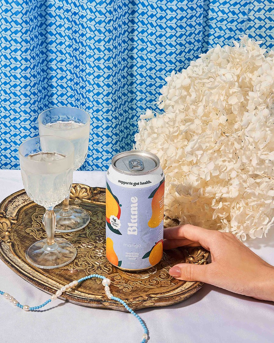 a hand holding a blue kombucha with glasses and flowers in the background
