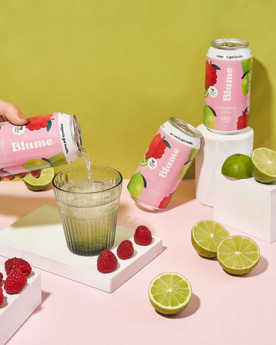 a hand pouring pink kombucha in a green glass with raspberries and fresh limes surrounding it