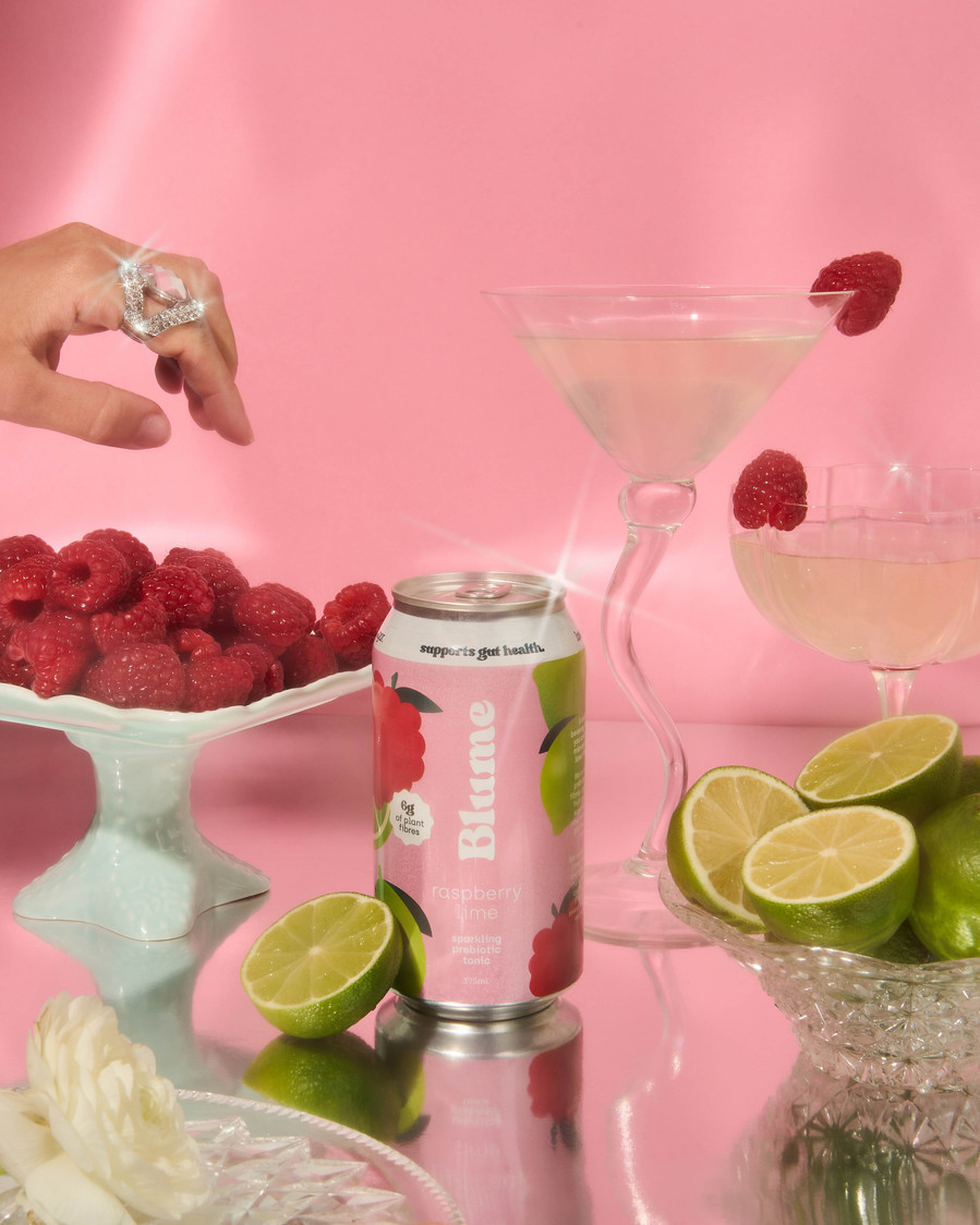 a hand with a big ring reaching for a pink kombucha can surrounded by limes and raspberries