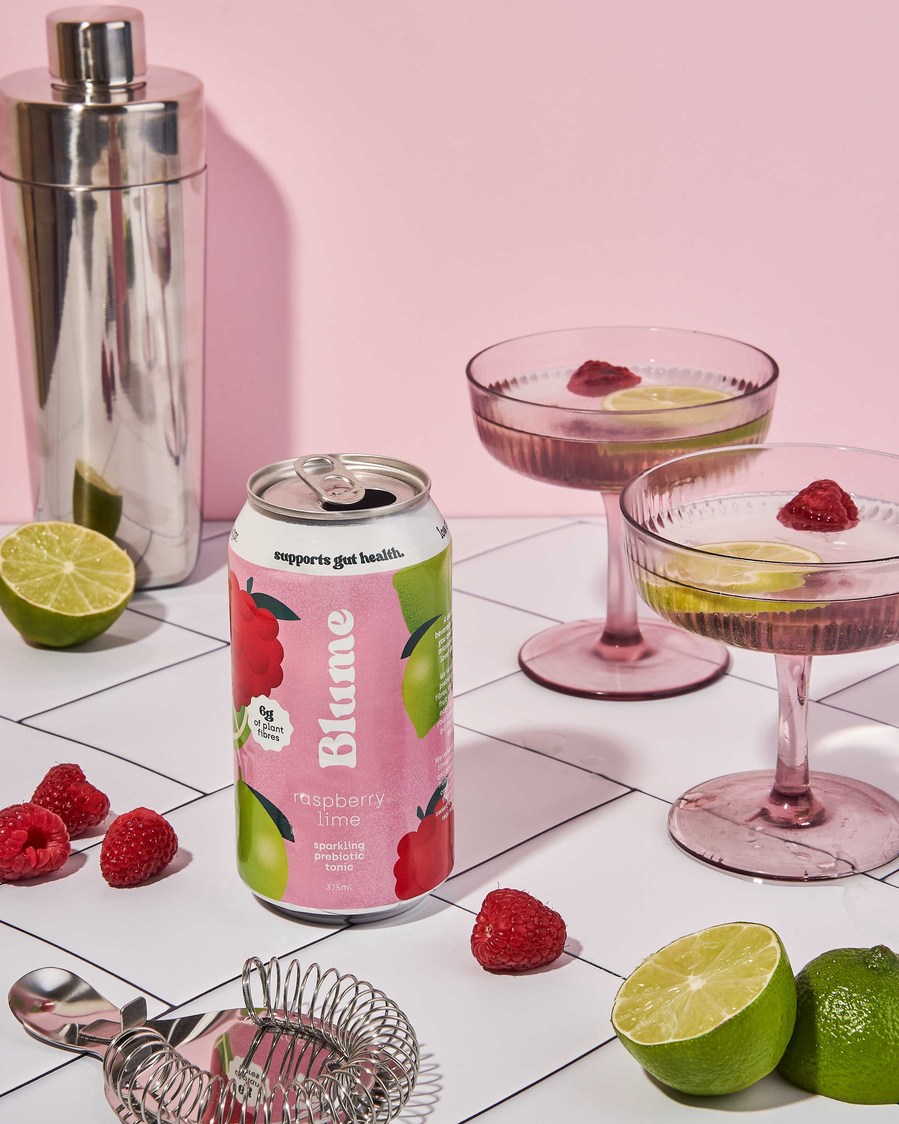 kombucha can on a table surrounded by pink glasses, raspberries and limes and a cocktail shaker