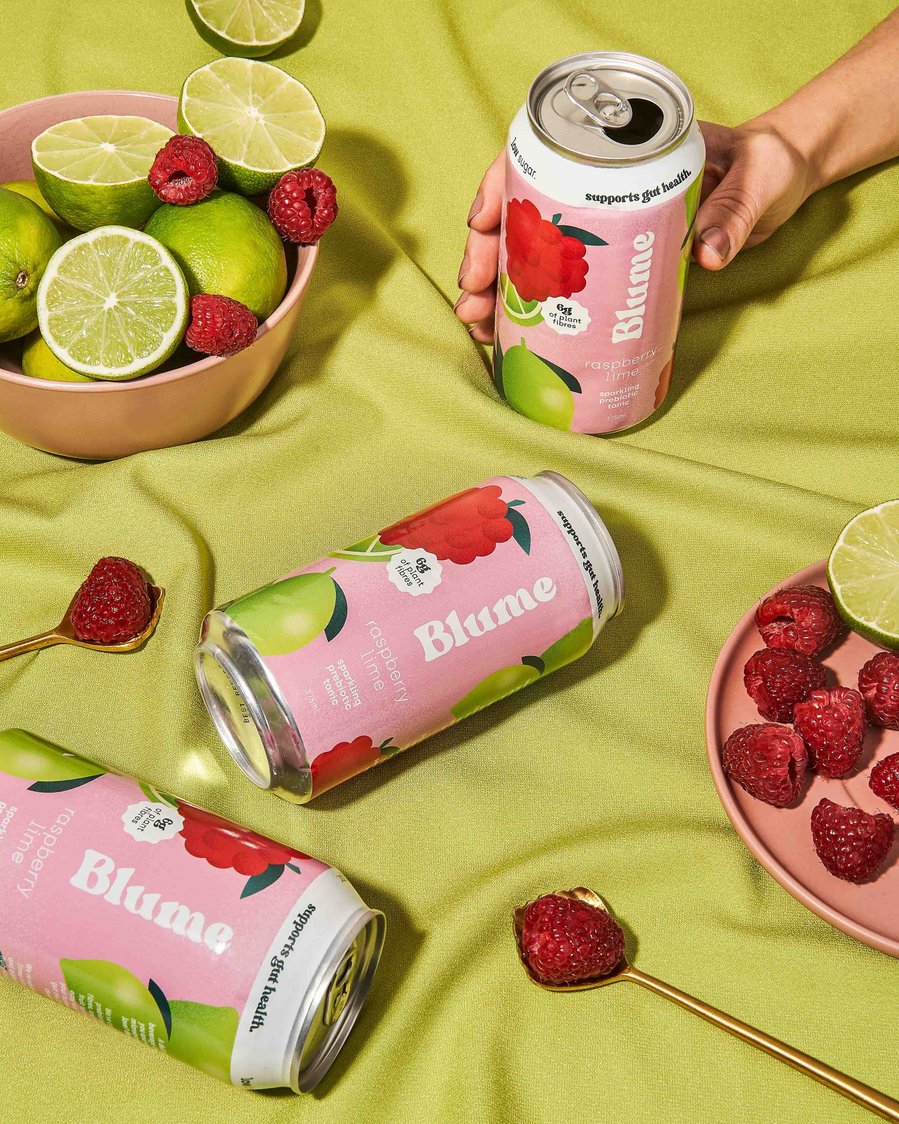 three kombucha cans with raspberries and limes lying around with a hand holding one of the kombuchas