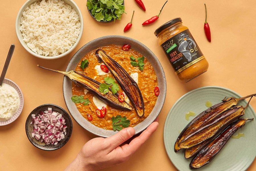 a colourful spread of Indian curry with eggplant and fresh ingredients lying around with a hand holding a plate with curry