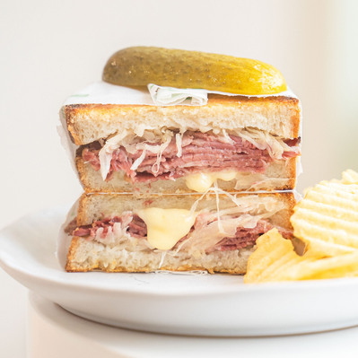 a big sandwich stack with ham, cheese, sauerkraut with a pickle on top and crisps on the side