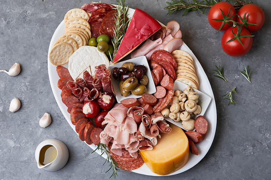 “Hearty charcuterie board with cold cuts, cheeses, olives, and crackers surrounded by garlic and tomatoes