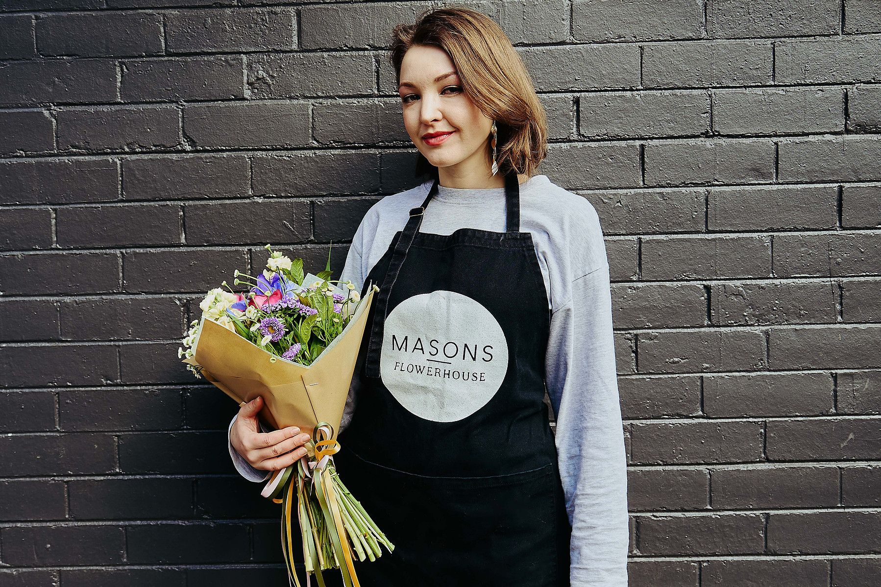 Florist wearing apron holding bouquet, standing against dark brick wall — Melbourne portrait and small business photography