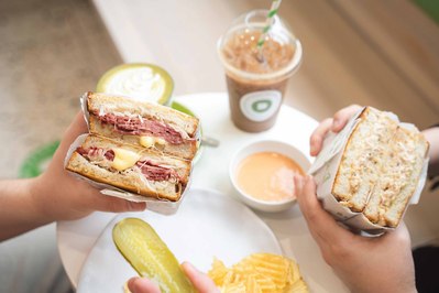 hands holding big sandwiches cut in half with iced drinks on the table