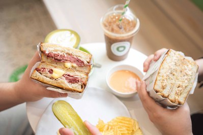 hands holding big sandwiches cut in half with iced drinks on the table