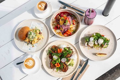 a flatlay of brunch items such as zucchini fritters, avocado on toast, chilli scramble and pancakes on a white table in a cafe