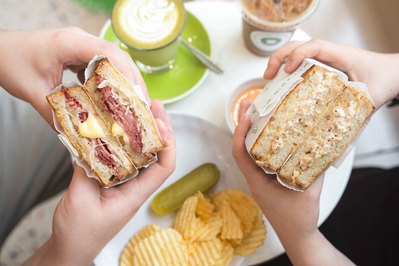 hands holding big sandwiches with coffees sitting on the table