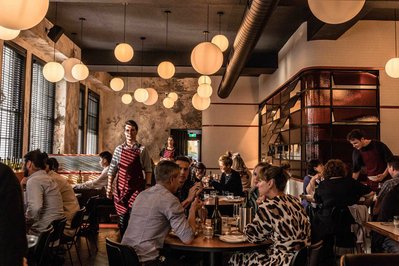 a busy modern restaurant with people sitting at tables and a waiter walking past with afternoon light coming through tall windows