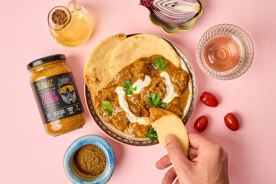 a colourful spread of Indian curry dinner with a hand dipping naan bread in a curry on a pink tablecloth