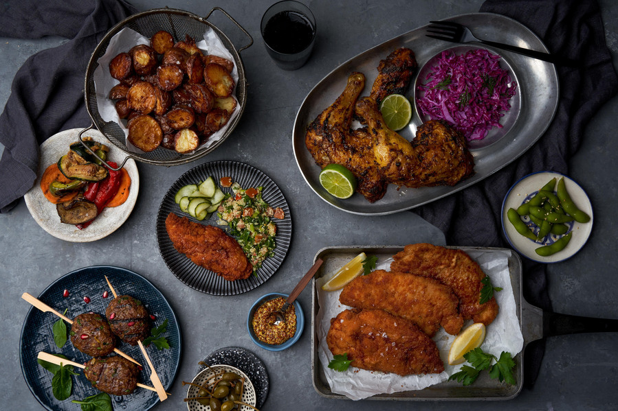 Dinner spread on the dark table containing of chicken schnitzels, chicken thighs, roasted potatoes and salad