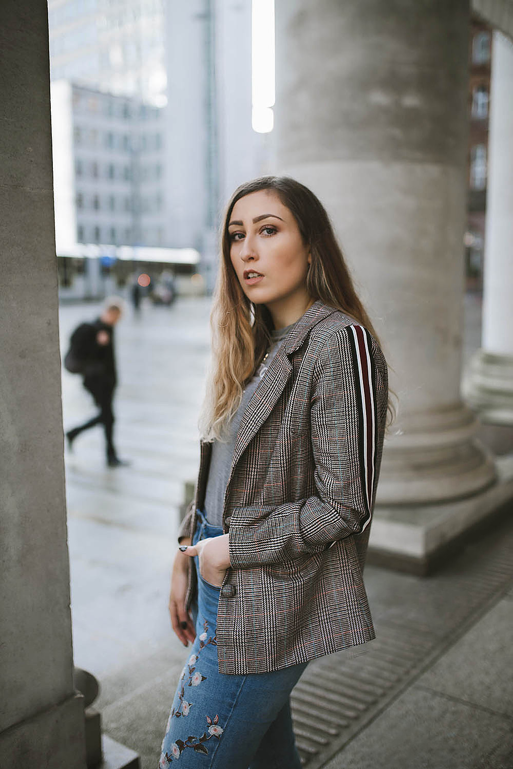 girl stands within Manchester city centre wearing floral jeans, and a blazer jacket with long brown hair