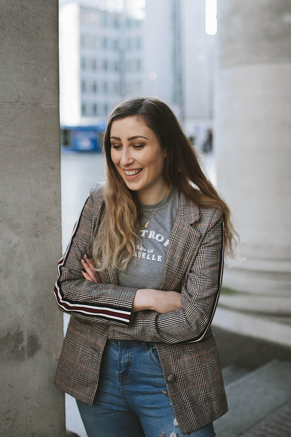 girl stands smiling within Manchester city centre wearing floral jeans, and a blazer jacket with long brown hair