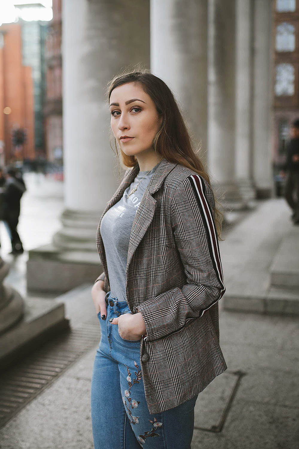 girl stands within Manchester city centre wearing floral jeans, and a blazer jacket with long brown hair