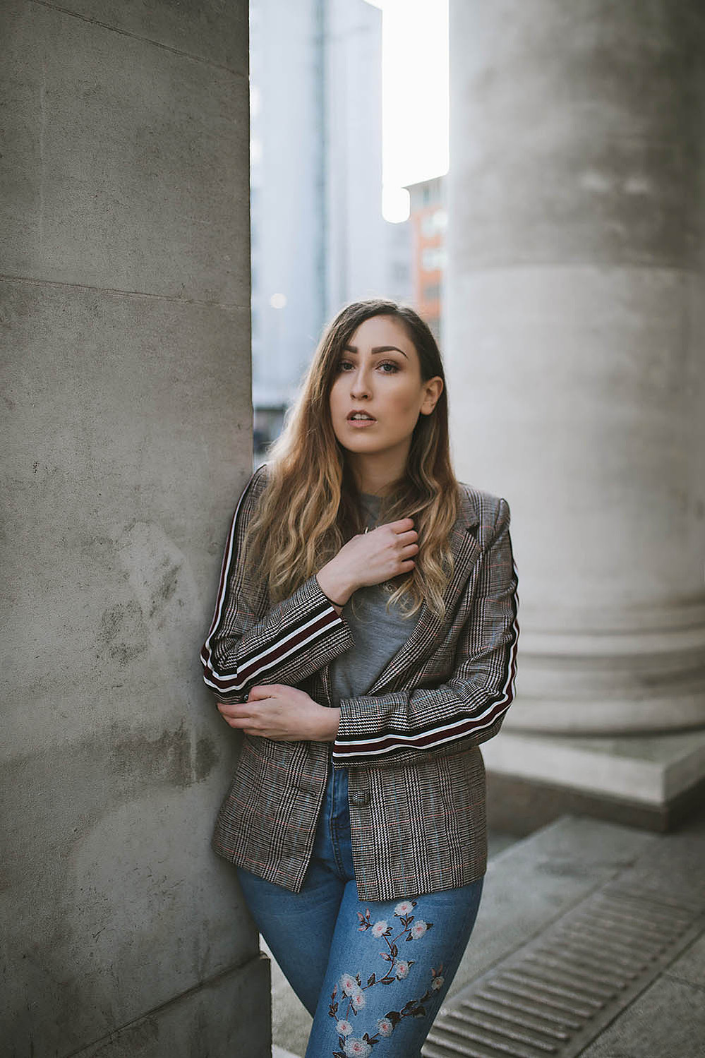 girl stands within Manchester city centre wearing floral jeans, and a blazer jacket with long brown hair