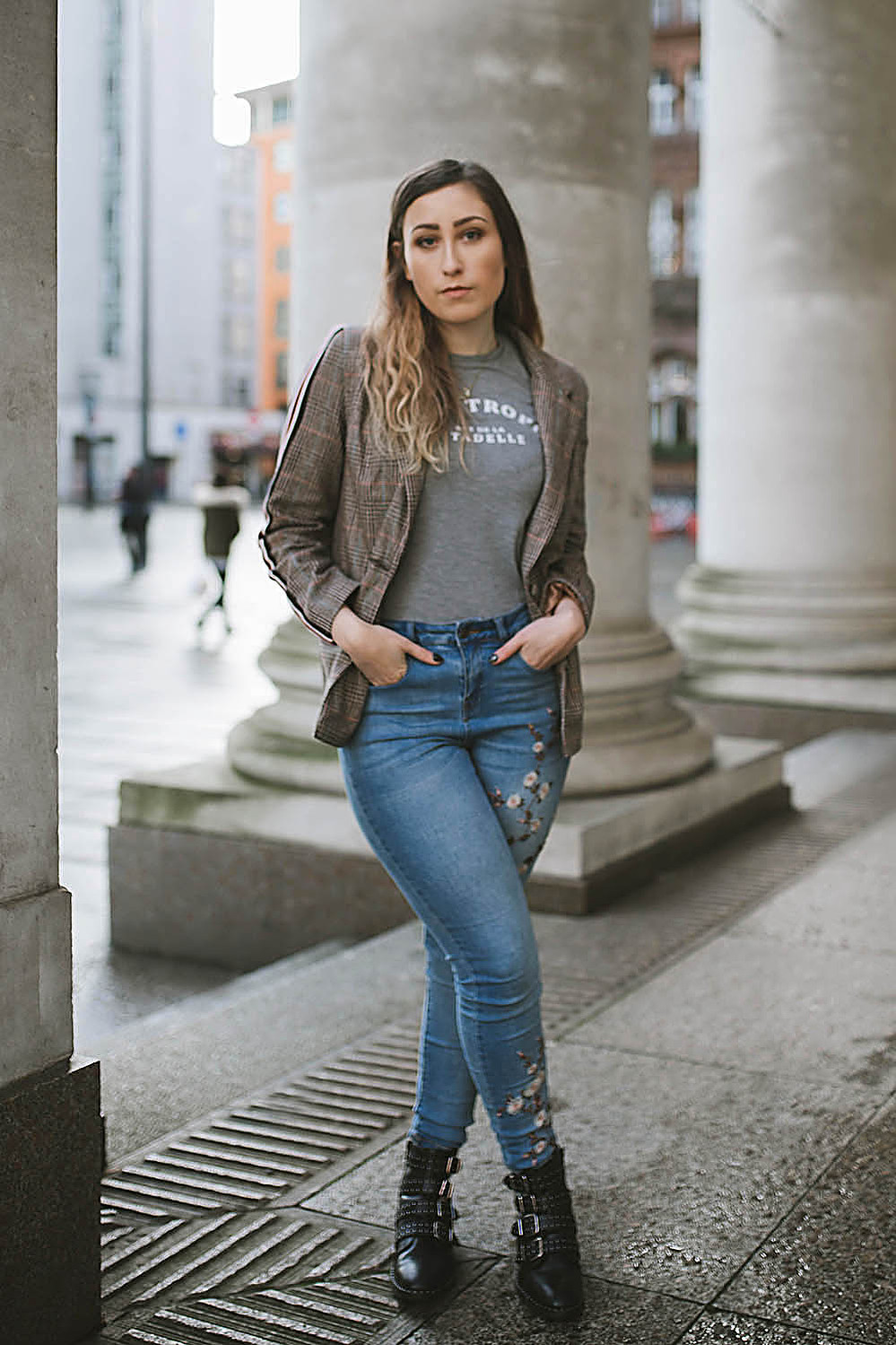 girl stands within Manchester city centre wearing floral jeans, and a blazer jacket with long brown hair