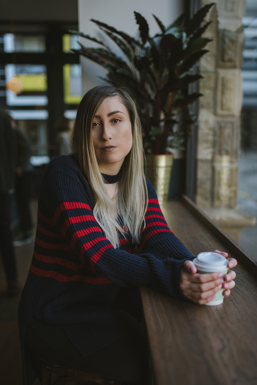 girl sits in coffee bar wearing striped jumper dress, with brown to blonde balayage hair nursing a coffee in a takeaway cup